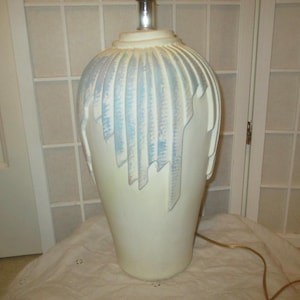 May include: A large, off-white ceramic table lamp with a bulbous base and a silver-toned metal top. The lamp features a decorative design of vertical, light blue accents. The lamp is on a white surface.