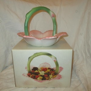 May include: A decorative ceramic basket with a pink and white flower-shaped bowl and a green handle. The basket sits atop a box with a similar floral design. The handle is green, and the bowl has a pink interior.