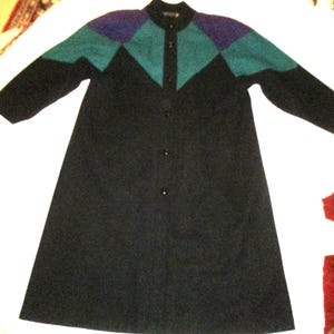 May include: A black long coat with a geometric color block design on the shoulders and upper chest. The color block features teal, purple, and black diamond shapes. The coat has a button-up front and long sleeves.