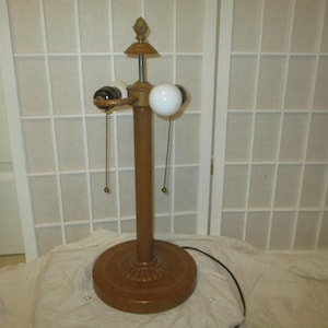 Peut inclure: Une lampe de table en bois brun avec trois ampoules et des chaînes de tirage. La lampe a un fleuron décoratif sur le dessus et une base ronde.