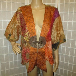 May include: An open-front, patchwork kimono-style jacket in shades of orange, gold, and pink. The jacket features a decorative clasp and a flowing design. The sleeves are short and wide. The garment is displayed on a mannequin.