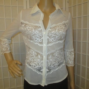May include: A white button-down shirt featuring sheer fabric panels and intricate floral lace detailing across the chest and sleeves. The garment has a classic collar, three-quarter length sleeves with lace cuffs, and a vertical button placket.