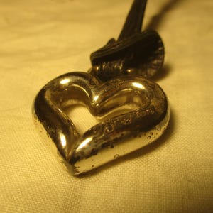 May include: A silver heart-shaped pendant with a black leather strap. The heart has a textured surface and a hollow centre. The pendant is likely made of metal and is displayed against a light-coloured background.