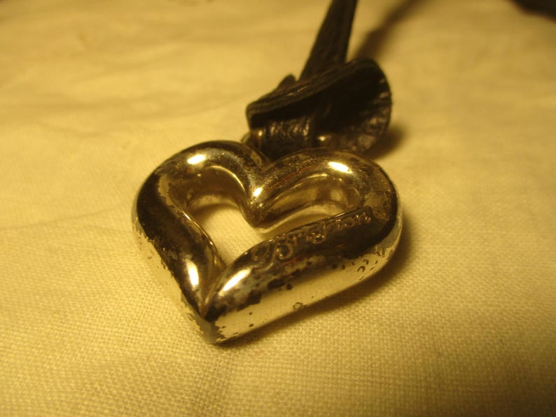 May include: A silver heart-shaped pendant with a black leather strap. The heart has a textured surface and a hollow centre. The pendant is likely made of metal and is displayed against a light-coloured background.