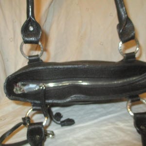 May include: Black leather tote bag with silver-tone metal detailing. The bag has a zip closure and two handles attached with metal rings. The interior is visible. A practical and stylish accessory for daily use.