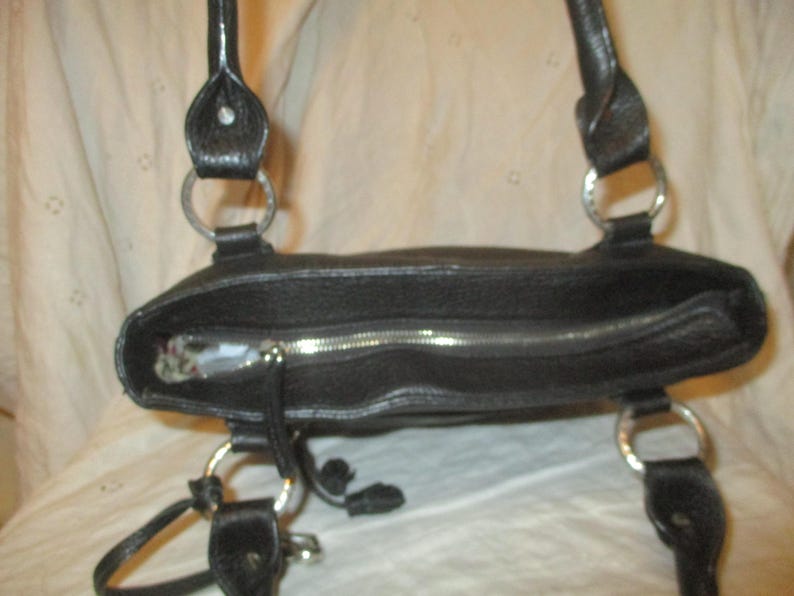 May include: Black leather tote bag with silver-tone metal detailing. The bag has a zip closure and two handles attached with metal rings. The interior is visible. A practical and stylish accessory for daily use.