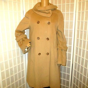 Puede incluir: Un abrigo beige de doble botonadura con cuello ancho y mangas largas. El abrigo tiene seis botones marrones y una falda acampanada. Las mangas tienen puños decorativos. El abrigo se exhibe en un maniquí.