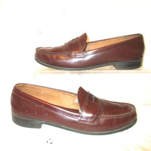 May include: A pair of burgundy leather penny loafers. The shoes are displayed on a white surface. The loafers have a classic design with a rounded toe and a low heel. The leather has a glossy finish.