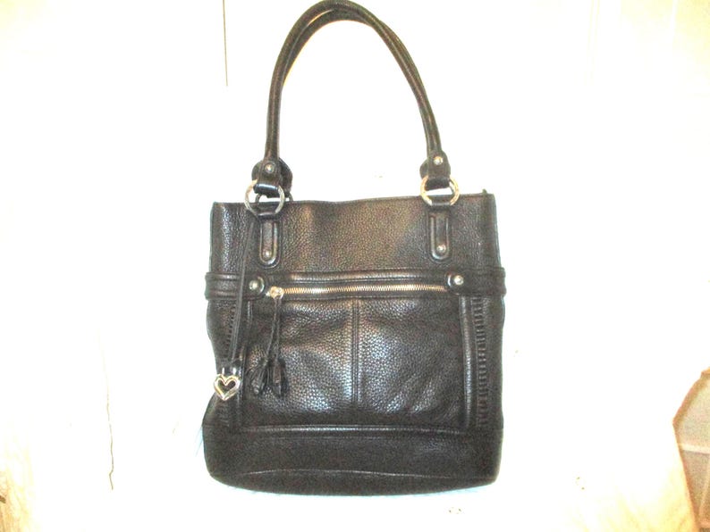 May include: Black leather tote bag with double handles and silver-tone hardware. Features a front zipped pocket, a decorative tassel, and a heart-shaped charm. The bag has a textured finish.