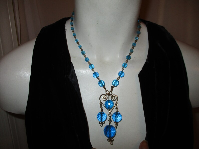 May include: A blue glass bead necklace with a dangling heart-shaped charm. The necklace is made of small, round beads and has a silver-toned metal chain.