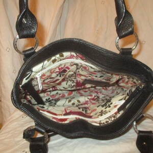 May include: Black leather tote bag with silver metal rings and handles. The interior has a floral pattern with red, pink, and white flowers on a cream background. The bag features a zipped closure.