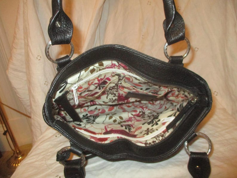 May include: Black leather tote bag with silver metal rings and handles. The interior has a floral pattern with red, pink, and white flowers on a cream background. The bag features a zipped closure.