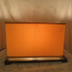 May include: A rectangular table lamp featuring a warm orange fabric shade that emits a soft glow. The lamp has a dark brown wooden base with a flat, rectangular design and a black power cord extending from the side of the base.