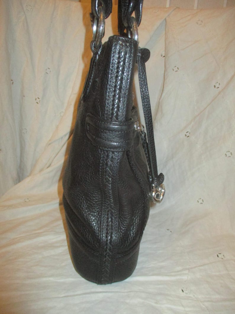 May include: Black leather handbag with silver-tone metal hardware. The bag has a textured surface and a braided detail along the side. The handles are connected with large silver rings. A small strap with a silver charm hangs from the side.