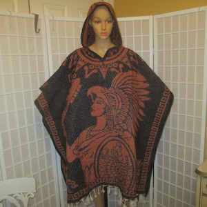 May include: A hooded poncho featuring a dark brown and burnt orange woven pattern. The design displays a profile of a figure wearing an elaborate feathered headdress and holding a circular shield. The hem is finished with white fringe tassels.