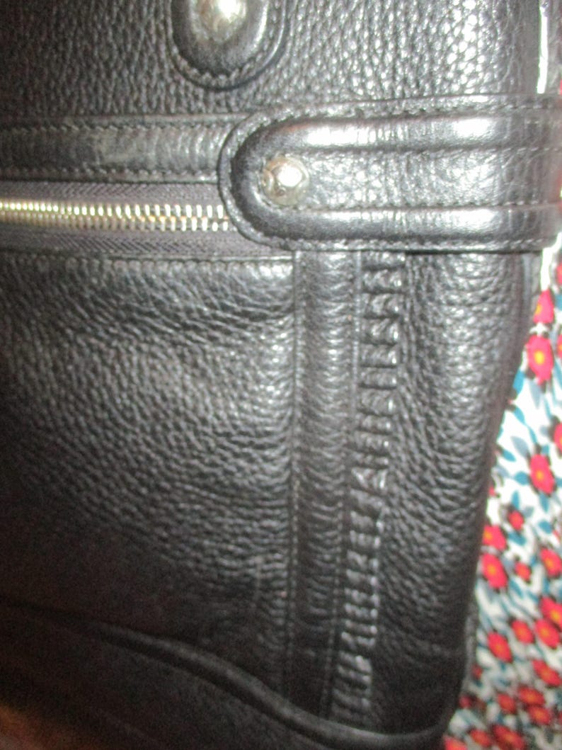 May include: Close-up of a black leather bag with a textured surface. The bag features a silver zip, decorative stitching, and silver rivets. The leather appears to be high-quality, with visible grain and texture.