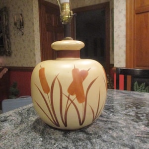 Puede incluir: Una lámpara de mesa de cerámica con una base beige y cañas pintadas de marrón. La lámpara tiene una pantalla marrón y beige.