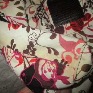 May include: Close-up of a floral patterned handbag interior. The fabric features a cream background with a repeating pattern of pink, red, and brown flowers and leaves. A black leather accent is visible.