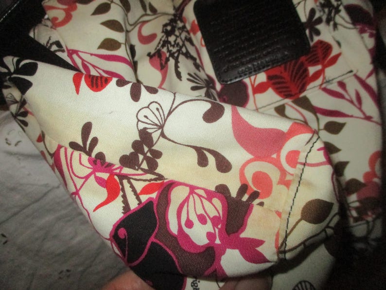 May include: Close-up of a floral patterned handbag interior. The fabric features a cream background with a repeating pattern of pink, red, and brown flowers and leaves. A black leather accent is visible.