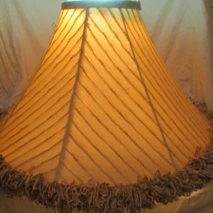 May include: A lampshade with a warm, yellow-orange hue, featuring a textured, diagonal striped pattern. The bottom edge is trimmed with a fringe. The lampshade is cone-shaped.
