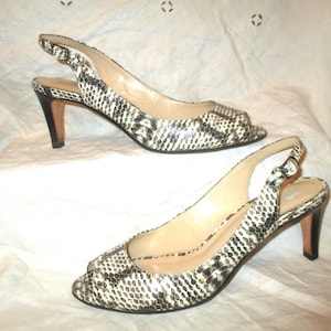 May include: A pair of open-toe slingback heels with a snakeskin pattern in black, white, and gray. The heels have a slender, dark brown heel and a buckle closure. The shoes are displayed on a white surface.