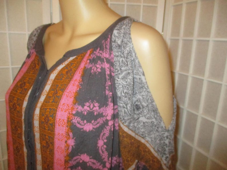 May include: A patterned top with a V-neckline and cut-out shoulders. The top features a mix of colors, including pink, orange, gray, and white. The fabric has a floral and geometric design.