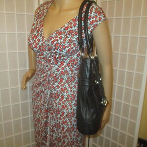 May include: A black leather handbag with silver-tone details is held by a mannequin. The bag has a structured shape with a top handle and a shoulder strap. The mannequin is wearing a floral print dress with red flowers.