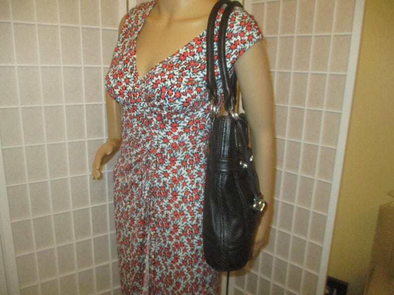 May include: A black leather handbag with silver-tone details is held by a mannequin. The bag has a structured shape with a top handle and a shoulder strap. The mannequin is wearing a floral print dress with red flowers.