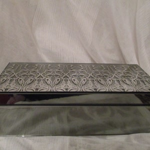 May include: A mirrored jewelry box with a decorative pattern on the lid. The box is rectangular and has a dark gray finish.