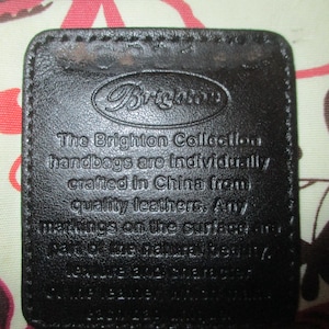 May include: A close-up of a dark brown leather tag with the Brighton logo and text. The tag is rectangular with rounded corners and stitched edges. The text reads "The Brighton Collection handbags are individually crafted in China from quality leathers."