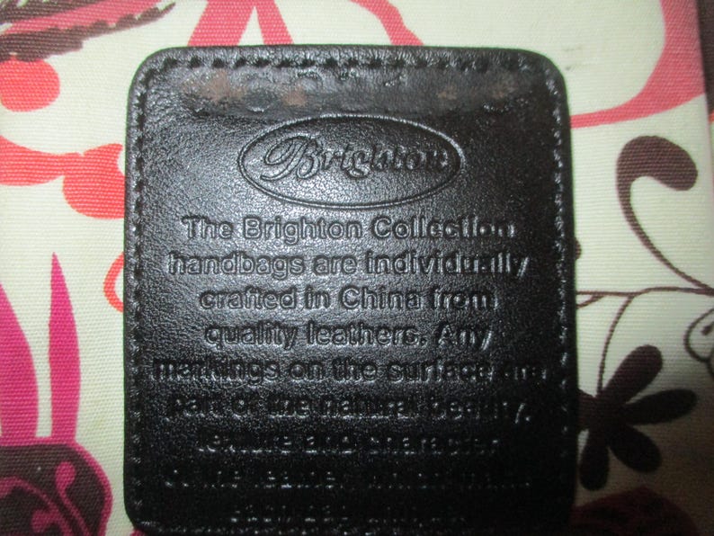May include: A close-up of a dark brown leather tag with the Brighton logo and text. The tag is rectangular with rounded corners and stitched edges. The text reads "The Brighton Collection handbags are individually crafted in China from quality leathers."