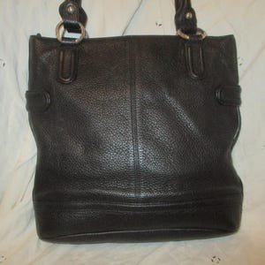 May include: Black leather tote bag with double handles and silver-tone metal rings. The bag has a textured surface and a rectangular shape with a structured design. Features include a top handle and side buckle details.