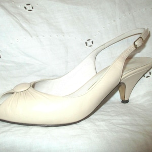 May include: Cream-colored slingback sandal with a peep-toe design. The shoe features a small, curved heel and an adjustable strap with a buckle. The sandal has a rounded toe and a decorative button detail on the vamp.