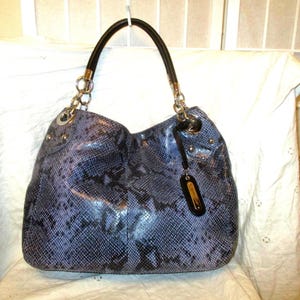 May include: A blue and black snakeskin-patterned handbag with a black handle and silver chain straps. The bag has a rounded shape and a small black tag with gold accents. The bag is sitting on a white surface.