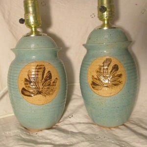 May include: Pair of ceramic table lamps. Each lamp has a light blue speckled glaze, a round tan medallion with a brown leaf design, and a brass lamp holder. The lamps are approximately 12 inches tall.