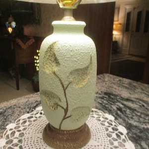 Peut inclure: Une lampe de table en céramique avec une base vert clair et des accents de feuilles d'or. La base a une surface texturée et présente une branche stylisée avec des feuilles.