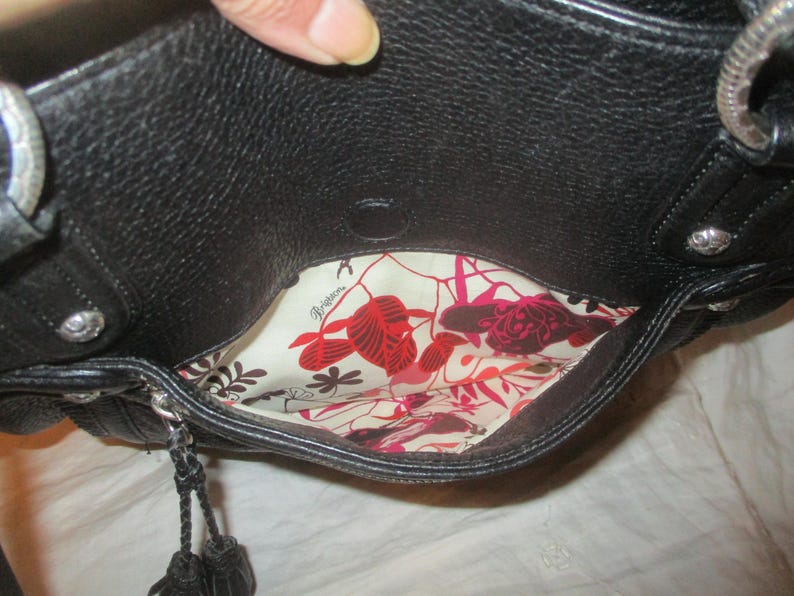 May include: Black leather handbag with silver-tone detailing. The interior has a floral print lining in red, pink, and cream. The bag features a top handle and a decorative tassel.
