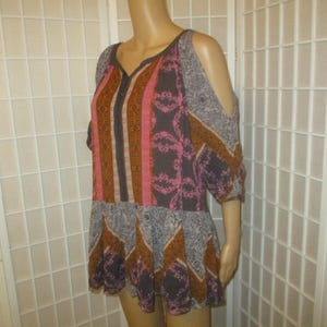 May include: A patterned, open-shoulder top in shades of pink, brown, and gray. The top features a V-neck and a flared hem. The sleeves are short and have cut-out details at the shoulders. The fabric has a mix of geometric and floral patterns.