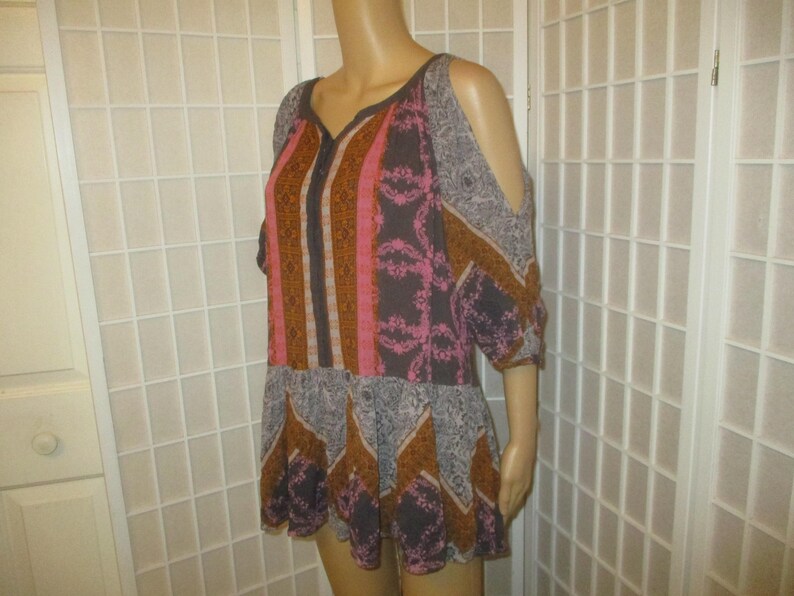 May include: A patterned, open-shoulder top in shades of pink, brown, and gray. The top features a V-neck and a flared hem. The sleeves are short and have cut-out details at the shoulders. The fabric has a mix of geometric and floral patterns.