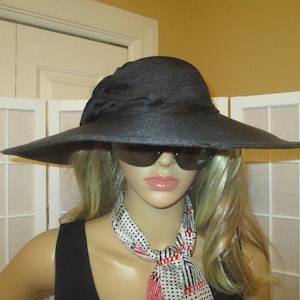 Puede incluir: Un sombrero negro de ala ancha con un gran lazo en la parte delantera. El sombrero lo lleva una persona con el pelo rubio y gafas de sol. La persona lleva un top negro de tirantes y una bufanda de lunares blancos y rojos.