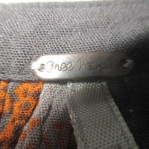 May include: Close-up of a gray fabric with an orange and black pattern. A metal tag with the words "Free People" is attached to the fabric. The tag is oval-shaped and has a dark, metallic finish.