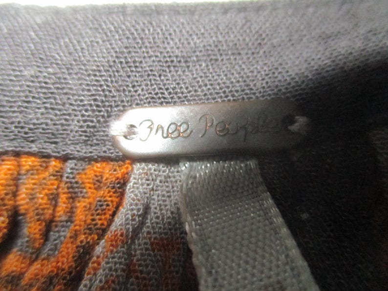 May include: Close-up of a gray fabric with an orange and black pattern. A metal tag with the words "Free People" is attached to the fabric. The tag is oval-shaped and has a dark, metallic finish.