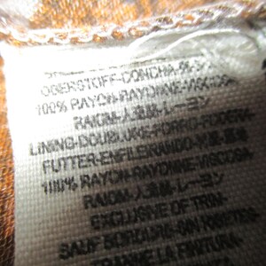 May include: Close-up of a white fabric tag with black text. The tag includes the text "100% RAYON RAYONNE-VISCOSA" and other text in multiple languages. The tag is attached to a brown and orange patterned fabric.
