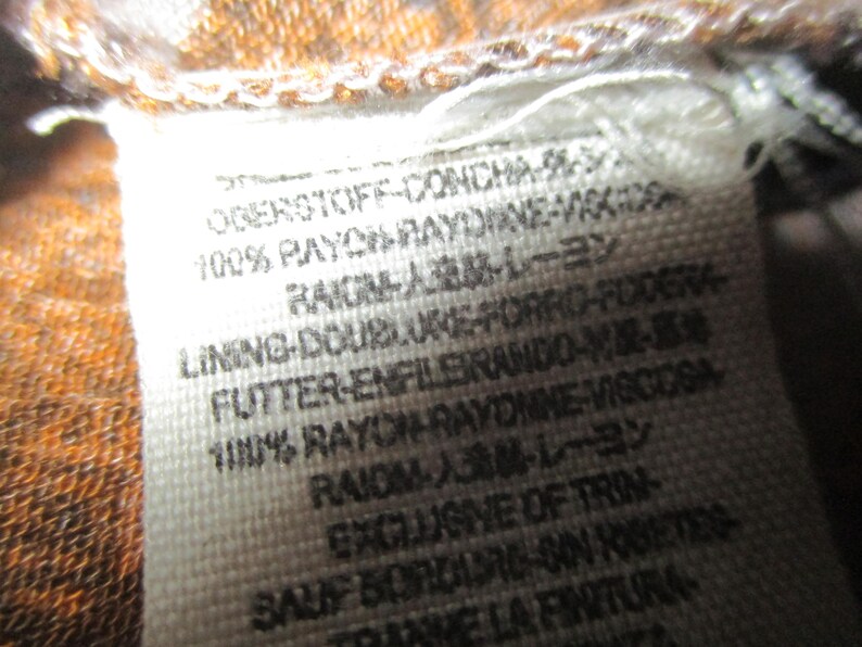 May include: Close-up of a white fabric tag with black text. The tag includes the text "100% RAYON RAYONNE-VISCOSA" and other text in multiple languages. The tag is attached to a brown and orange patterned fabric.