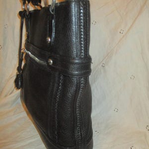 May include: Black leather tote bag with silver-tone metal detailing. The bag has a textured finish, a zip closure, and double handles. It features a decorative strap with silver studs and a small tassel. A classic, stylish design.