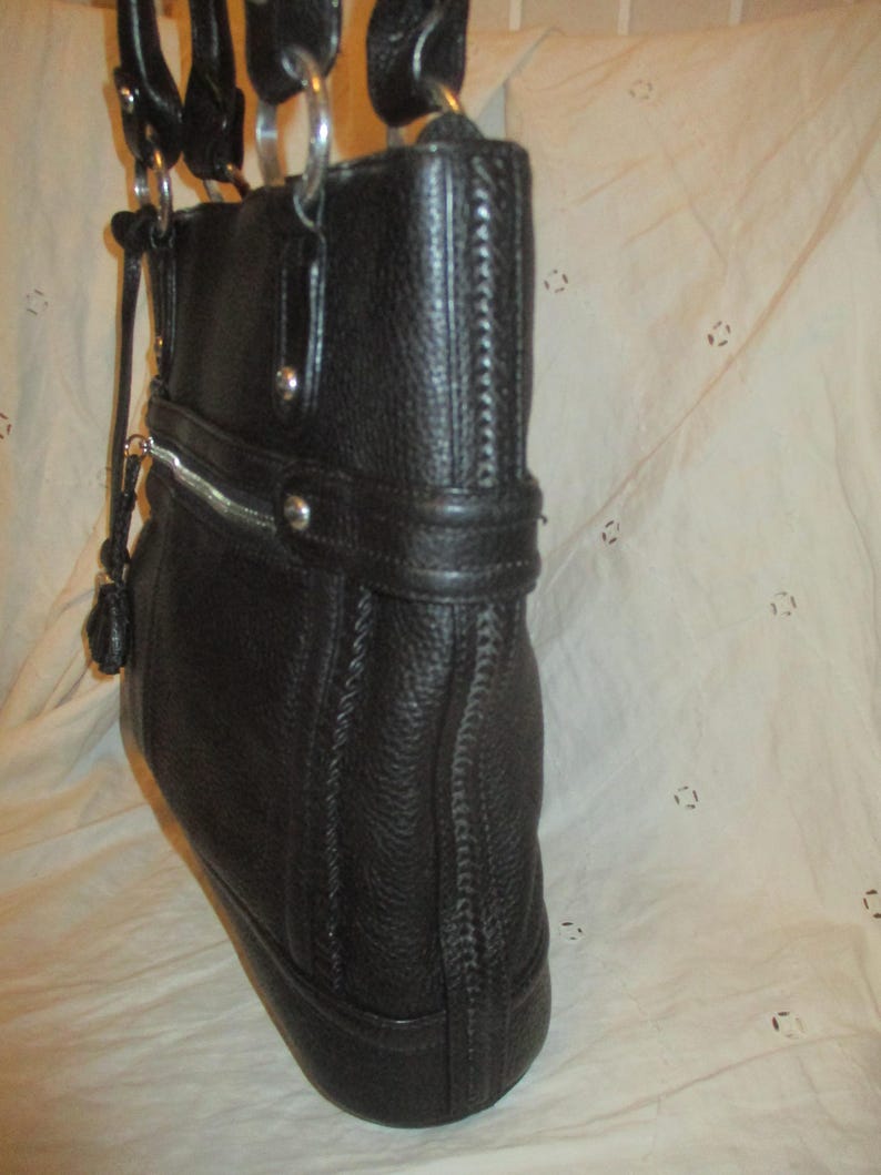 May include: Black leather tote bag with silver-tone metal detailing. The bag has a textured finish, a zip closure, and double handles. It features a decorative strap with silver studs and a small tassel. A classic, stylish design.