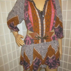 May include: A patterned tunic dress in shades of gray, brown, pink, and orange. The dress features a V-neckline, three-quarter sleeves, and a cinched waist with a brown belt and decorative buckle. The dress has a flowing skirt.