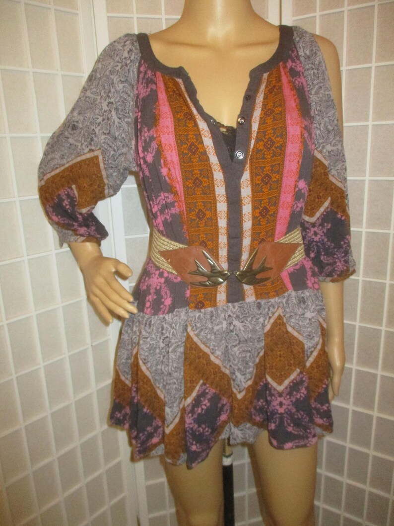 May include: A patterned tunic dress in shades of gray, brown, pink, and orange. The dress features a V-neckline, three-quarter sleeves, and a cinched waist with a brown belt and decorative buckle. The dress has a flowing skirt.