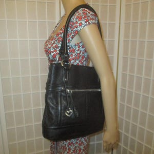 May include: Black leather tote bag with a textured finish. The bag has a silver zip, a heart-shaped charm, and a shoulder strap. The bag is held by a person wearing a floral print dress.