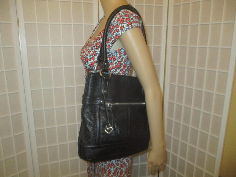 May include: Black leather tote bag with a textured finish. The bag has a silver zip, a heart-shaped charm, and a shoulder strap. The bag is held by a person wearing a floral print dress.